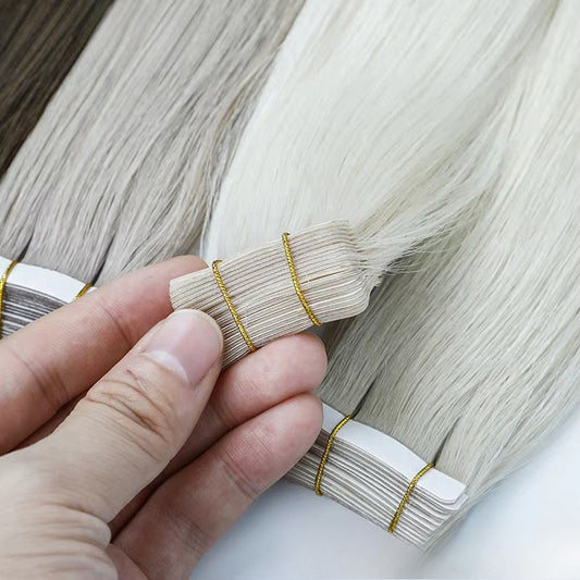 Close-up of light blonde tape-in hair extensions, held by a hand to show the adhesive tape and hair texture.