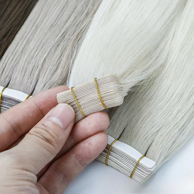 Close-up of light blonde tape-in hair extensions, held by a hand to show the adhesive tape and hair texture.
